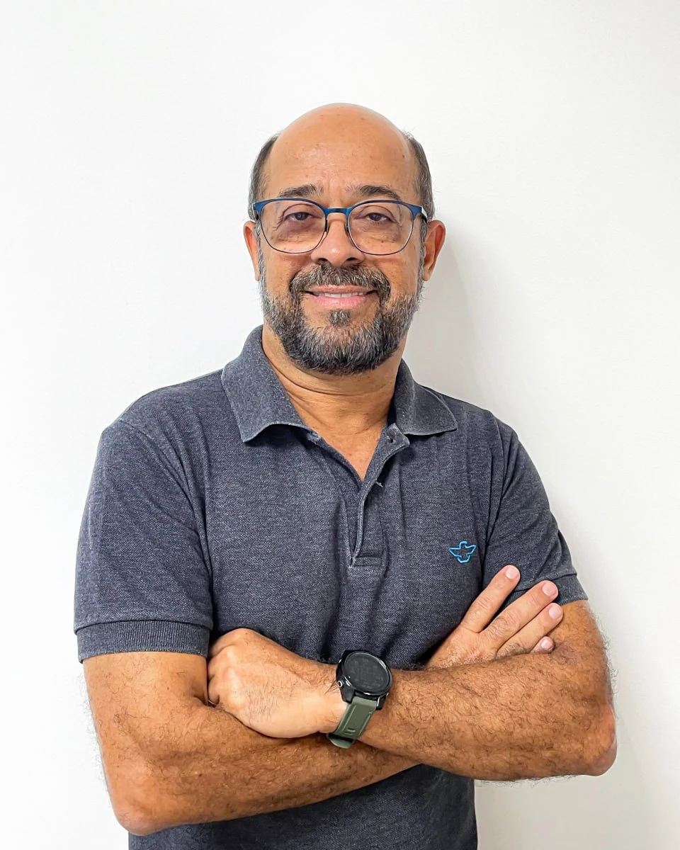 Retrato de Lula Moura, de braços cruzados, usando óculos e camisa polo cinza.